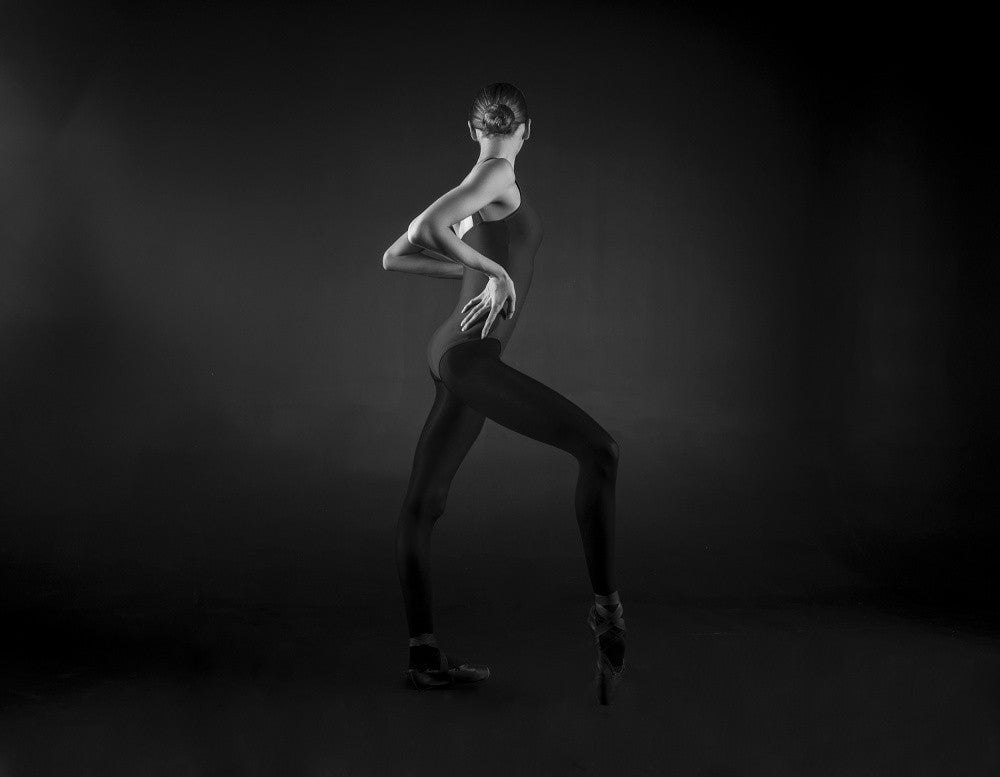 ballerina in a blue leotard on a dark background stands in a closed pose immersed in thought