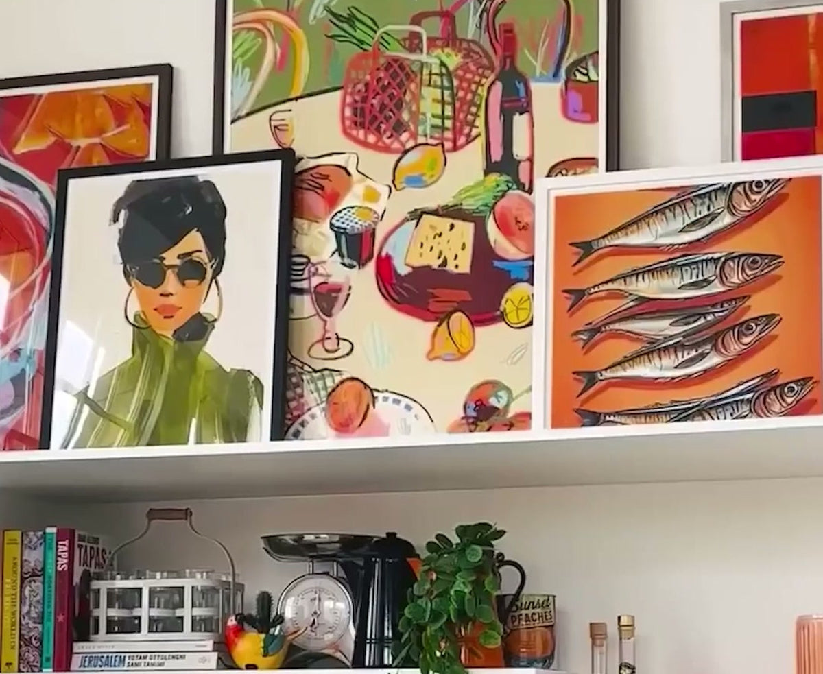 Shelf of framed prints including a portrait in sunglasses and sardine illustrations in a colourful living room