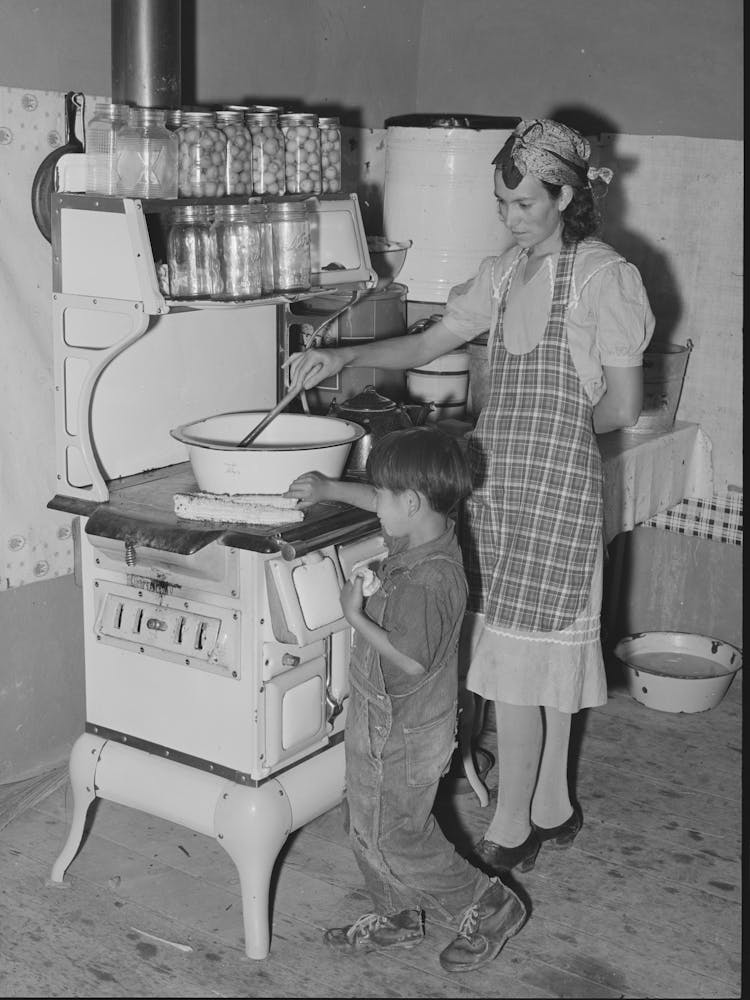Spanish American Woman Stirring Pan Of Cooking Beans, Small Boy Is Roasting Sweet Corn On Top Of The Stove