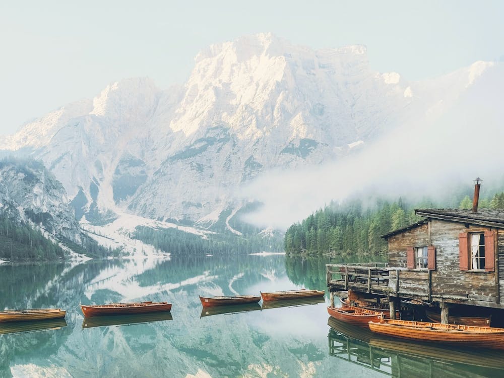 Dolomites, Italy I Lago di Braies lake with its wooden cabin and boats in front of the snow mountain from morning mist fog to the moody aesthetic atmosphere photography of sunrise pastel light in misty forest