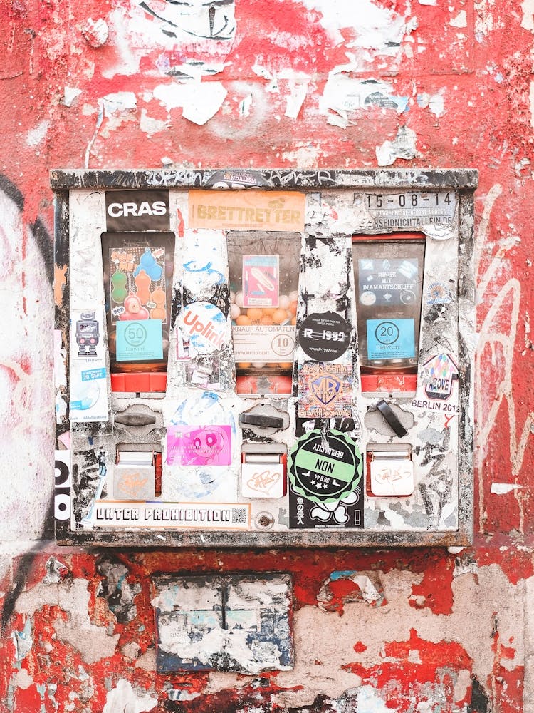 Berlin, Deutschland I Süßigkeitenautomat voller Sticker an beschädigter roter Wand in rauer Straßenszene mit groben Texturen und Underground-Berliner-Vibes Ästhetik für Liebhaber urbaner Kunst, edgy Fotografie