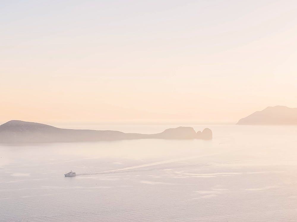 Milos, Greece I Summer pastel aesthetic sunset over the mountains and a boat on Mediterranean sea for a minimalist fine art photography of soft soothing calming nature in pink orange retro tones
