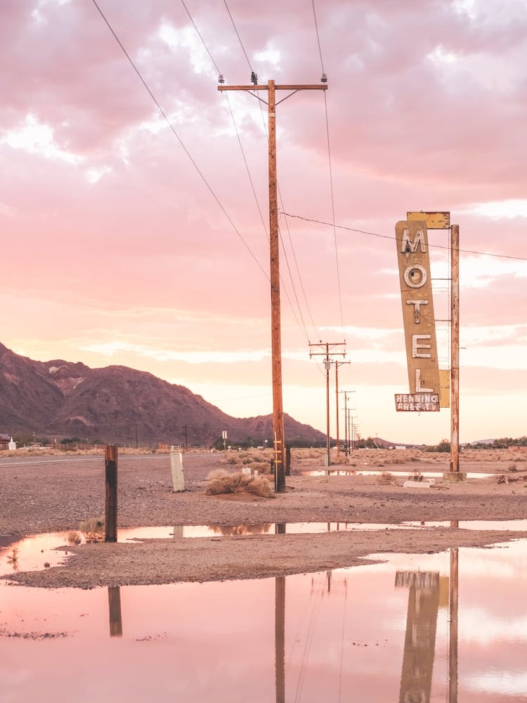 Route 66, USA I Sunset in front of a vintage retro motel neon sign in a California desert landscape to pastel pink sky sun aesthetic and a mirrored reflection in water photography in front of the Bagdad Cafe
