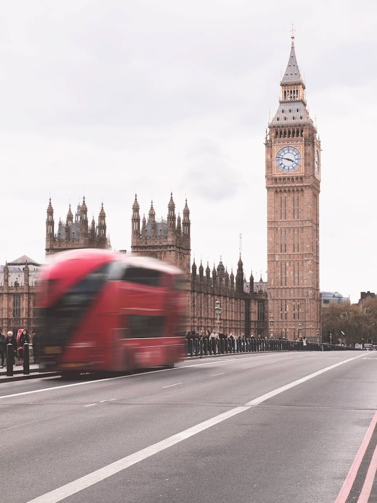 London, England I Big Ben clock tower and a british iconic red bus in motion on Westminster Bridge under an aesthetic autumn moody cloudy grey sky on the road and geometric london brick architecture photography