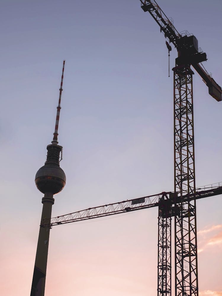 Berlin, Germany I Fernsehturm tower silhouette and cranes in pastel pink and blue sunset sky light for a minimal industrial cityscape photography with golden hour glow and geometric urban composition
